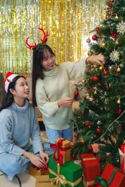 İki arkadaş bir Noel ağacını süslediler, gülümsüyorlar ve Noel Baba şapkaları ve süslemelerle eğleniyorlar..