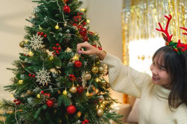 Noel Baba şapkalı genç bir kadın güzel dekore edilmiş bir Noel ağacına süs asmak için uzanır, ışıltılı bir arka planda bayram heyecanıyla gülümser.