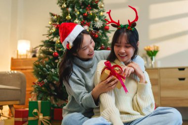 İki arkadaş Noel ağacının yanında neşeli bir anı paylaşıyor, biri hediye paketini tutuyor, diğeri de bir fincan. Şenlikli başlıkları çekicilik katıyor, sıcaklığı vurguluyor.