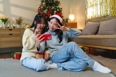 Tatil başlıklı iki arkadaş telefona bakıp gülümserken süslü bir Noel ağacının yanında oturuyorlar. Bayram heyecanı ve neşeli paylaşımı yansıtıyor..