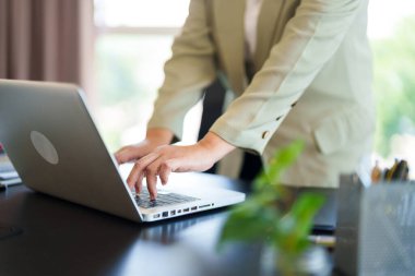 Masasında duran bir iş kadını, bilgisayarının üzerine yaslanmış, parlak, modern bir ofis alanında. Meşgul ve üretken bir ortamda çalışmaya hazır görünüyor..