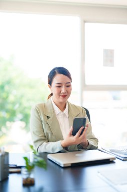 Hafif takım elbiseli gülümseyen bir iş kadını masasında oturuyor, telefonunu parlak bir ofis ortamında tutuyor, meşgul ve bağlı görünüyor..