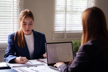 Takım elbiseli iki iş kadını bir masada çalışıyor, ikisi de bilgisayarlarına ve belgelerine odaklanmış. Profesyonel ofis ortamındalar.