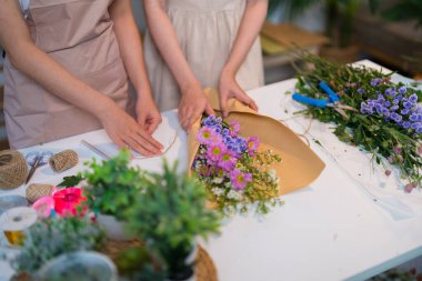 İki çiçekçi, parlak bir dükkanda saksı bitkileri ve renkli çiçeklerle çevrili güzel bir çiçek aranjmanı yapmaya odaklanmış..