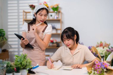 İki çiçekçi neşeyle bir çiçekçide iş birliği yapıyor, biri tablet tutarken diğeri ise canlı çiçeklerin arasında gülüyor..