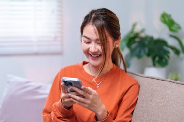 Genç bir kadın rahat bir kanepede rahatlar, modern bir oturma odasında akıllı telefonunu karıştırırken gülümser..