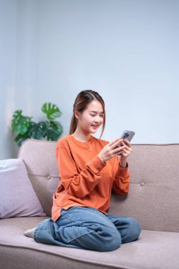 Genç bir kadın rahat bir kanepede rahatlar, modern bir oturma odasında akıllı telefonunu karıştırırken gülümser..