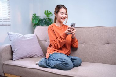 Genç bir kadın rahat bir kanepede rahatlar, modern bir oturma odasında akıllı telefonunu karıştırırken gülümser..