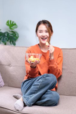 Kanepede oturan neşeli genç bir kadın TV izlerken kaseden atıştırmalıkların keyfini çıkarıyor..