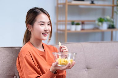 Kanepede oturan neşeli genç bir kadın TV izlerken kaseden atıştırmalıkların keyfini çıkarıyor..