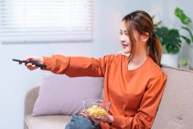 Kanepede rahat bir şekilde oturan genç bir kadın, elinde bir televizyon kumandası ve bir kase atıştırmalık, rahatlamış görünüyor..