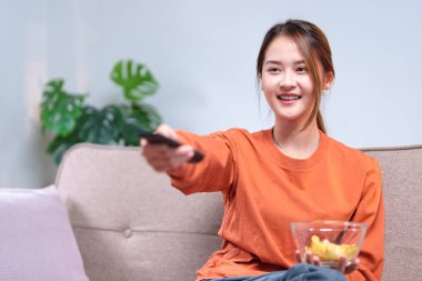 Kanepede rahat bir şekilde oturan genç bir kadın, elinde bir televizyon kumandası ve bir kase atıştırmalık, rahatlamış görünüyor..