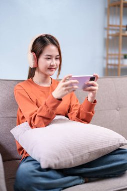 Kulaklık takan genç bir kadın, koltukta rahat bir şekilde otururken akıllı telefonuyla oyun oynuyor..