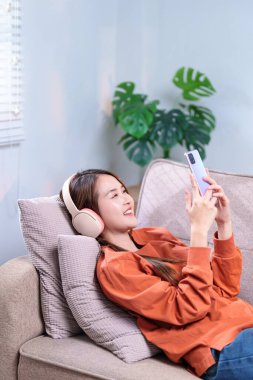 Rahat bir kadın kanepede uzanıyor, kablosuz kulaklıkla müzik dinliyor ve akıllı telefonunu tutuyor..