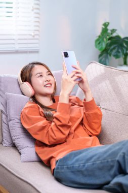 Rahat bir kadın kanepede uzanıyor, kablosuz kulaklıkla müzik dinliyor ve akıllı telefonunu tutuyor..