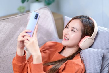 Rahat bir kadın kanepede uzanıyor, kablosuz kulaklıkla müzik dinliyor ve akıllı telefonunu tutuyor..