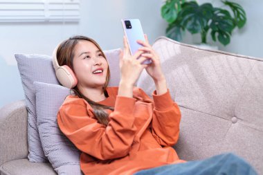 Rahat bir kadın kanepede uzanıyor, kablosuz kulaklıkla müzik dinliyor ve akıllı telefonunu tutuyor..