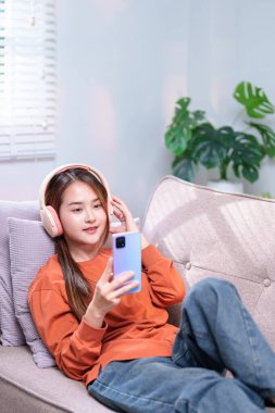 Kanepede oturan genç bir kadın, kablosuz kulaklıklarla, gülümsüyor ve akıllı telefondan en sevdiği melodileri dinliyor..