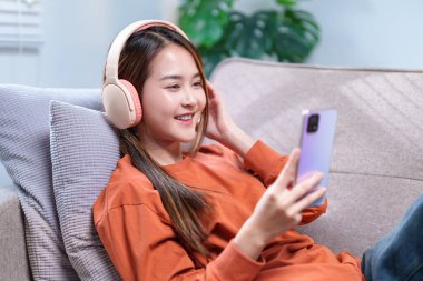 Kanepede oturan genç bir kadın, kablosuz kulaklıklarla, gülümsüyor ve akıllı telefondan en sevdiği melodileri dinliyor..