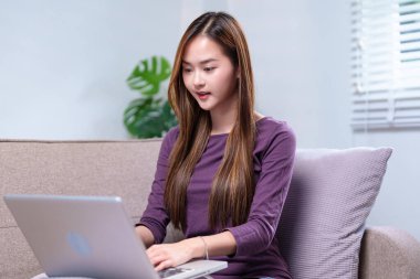 Dizüstü bilgisayarda çalışan genç bir kadın kanepede oturuyor, gülümsüyor ve odaklanmış görünüyor, rahat bir evde..