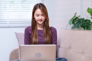 Dizüstü bilgisayarda çalışan genç bir kadın kanepede oturuyor, gülümsüyor ve odaklanmış görünüyor, rahat bir evde..