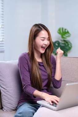 Dizüstü bilgisayar kullanırken heyecanla tezahürat yapan bir kadın, rahat bir oturma odasında başarı anını kutluyor..