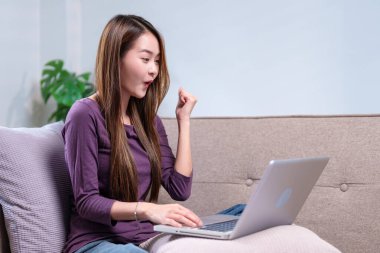 Dizüstü bilgisayar kullanırken heyecanla tezahürat yapan bir kadın, rahat bir oturma odasında başarı anını kutluyor..