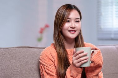 Elinde bir kupa kahve tutan genç bir kadın, kanepede sakin bir anın tadını çıkarıyor, rahat turuncu bir kazak giymiş, sakin bir ifadeyle.