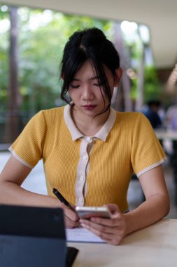 Genç Asyalı bir kadın üniversite kafesinde tabletle çalışırken akıllı telefon kullanıyor. Kitapları ve masada içkisi var.