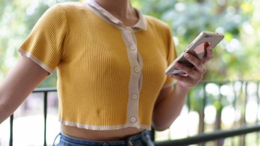 Üniversite ortamında doğal ışık alan bir yerde dururken akıllı telefon kullanan bir kadının ellerinin yakın plan fotoğrafı.