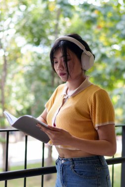 Genç Asyalı bir kadın, kablosuz kulaklık takıp deftere yazı yazıyor ve üniversite ortamında açık havada okuyor.