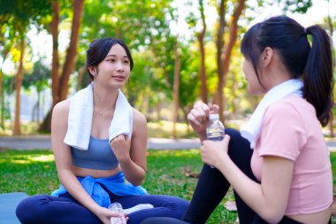 İki genç Asyalı kadın, yoga minderleri ve su şişeleriyle parkta egzersiz yaptıktan sonra dinlenip sohbet ediyorlar.