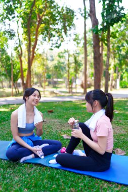 İki genç Asyalı kadın, yoga minderleri ve su şişeleriyle parkta egzersiz yaptıktan sonra dinlenip sohbet ediyorlar.