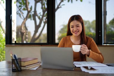 İş kadını kahve içerken doğal temalı bir ofiste dizüstü bilgisayarı ve masadaki belgelerle çalışıyor..