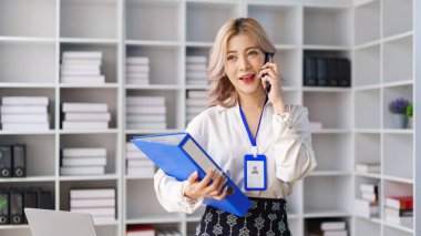 Modern ofiste proje dosyalarını tutarken akıllı telefondan konuşan profesyonel bir iş kadını.