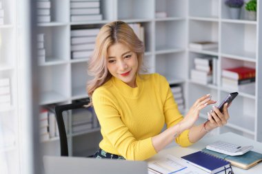 Ofis arasında bilgisayar ve defterle akıllı telefon kullanan gülümseyen bir iş kadını.