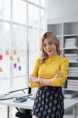Kendine güveni tam genç bir iş kadını modern ofis alanında dizüstü bilgisayar ve yapışkan notlarla çapraz duruyor.