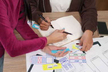 UI tasarım ekibindeki Asyalı kadınlar mobil uygulama arayüzü üzerinde işbirliği yapıyorlar. İşyerlerinde kullanıcı deneyimini (UX) geliştirmek için fikirleri bir deftere çiziyorlar..