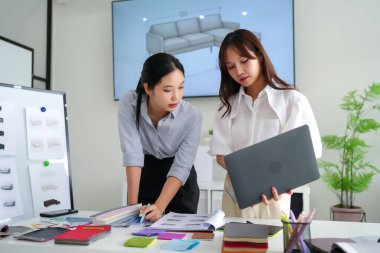 İki genç Asyalı kadın iç mimar kanepe tasarımında işbirliği yapıyorlar, kumaş örnekleri seçiyorlar, modern bir ofiste dizüstü bilgisayar kullanıyorlar. Renk paletleri ve koltuk çizimleriyle çalışma alanı.