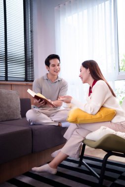 Yetişkin bir Asyalı çiftin kitap okuyup evde fikirleri tartışırken görüntüsü. Rahat bir oturma odasındaki kanepede dinlenerek boş vakitlerinin tadını çıkarıyorlar..