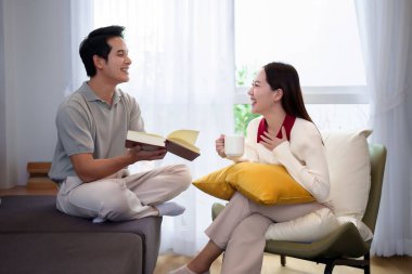 Asyalı yetişkin bir çiftin samimi oturma odalarında iç açıcı bir görüntüsünü yakalayın. Kadın kahvesini yudumlarken adam elinde bir kitap tutuyor. Neşeli bir bağ ve rahatlama anını paylaşıyorlar..