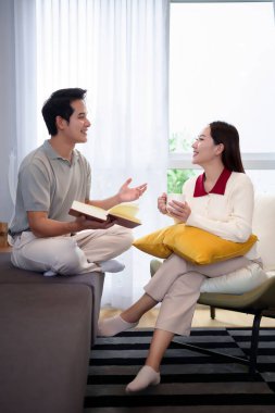 Mutlu bir Asyalı çift evde kaliteli zaman geçiriyor. Genç adam, yetişkin kadın bir fincan kahveyle bir sandalyede dinlenirken yüksek sesle bir kitap okuyor. Rahat bir yaşam alanında gülümsüyorlar ve konuşuyorlar..