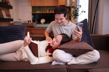 Genç bir Asyalı çift evdeki kanepelerinde kaliteli vakit geçirmekten zevk alır. Kadın kitap okurken, erkek cep telefonuyla bir şeyler paylaşır. Ev içinde günlük yaşam tarzı.