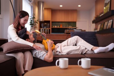 Rahat, yetişkin bir Asyalı çift evde müzik dinliyor. Kadın, erkeğin kanepede ukulele çalarken gülümsüyor. Oturma odasında samimi bir an, boş vakit ve ev hayatı sergiliyor.