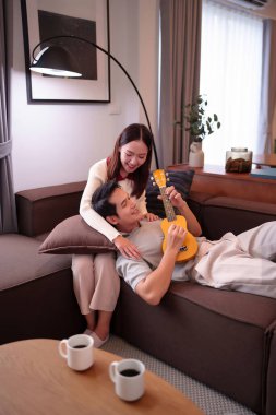 Genç bir Asyalı çift kanepede dinlenirken adam ukulele çalarken kadın ona sarılıyor. Kahve fincanlarıyla sıcak bir sahnede birlikte mutlu görünüyorlar.