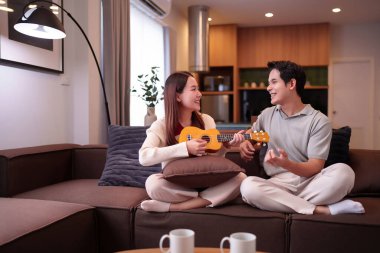 Mutlu bir Asyalı çift evde dinleniyor. Asyalı bir kadın ukulele çalıyor ve Asyalı bir adam onun rahat oturma odasında gülümsemesini dinliyor..