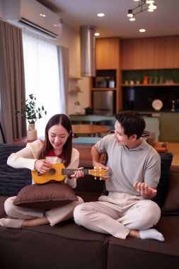 Yetişkin bir Asyalı erkek ve Asyalı genç bir kadının rahat oturma odalarında gitar çalıp müzik aletlerinin ve rahatlama seslerinin tadını çıkardıkları iç açıcı bir sahne.