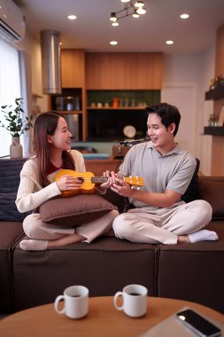 Asyalı çift evdeki kanepede birlikte ukulele oynuyor. Mutlu genç adam ve kadın gülümsüyor müzik dersinin keyfini çıkarıyor ilişki rahatlaması evde boş vakit geçiriyor..
