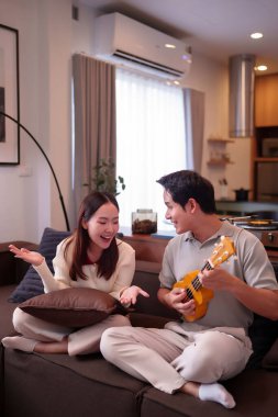Asyalı yetişkin bir çift kanepede gitar çalmaktan hoşlanır. Kadın gülerken erkek enstrüman çalıyor. Evde müzik aleti ukulele ile harika zaman geçiriyoruz..