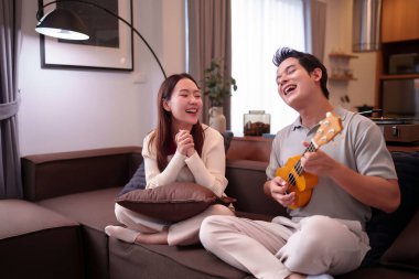 Yetişkin bir Asyalı çift rahat oturma odalarında ukulele müziğinin keyfini çıkarıyorlar. Birbirlerine bağlanıyorlar ve birlikte eğlenceli vakit geçiriyorlar. Güzel ilişkileri gülüşlerinde belli oluyor.